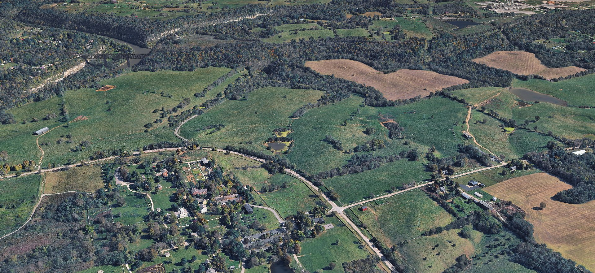 3D aerial view of the Shaker Village of Pleasant Hill historic core surrounded by rolling Mercer County farmland, with the Kentucky River palisades and High Bridge in the upper left and the E.W. Brown power station visible in the upper right.