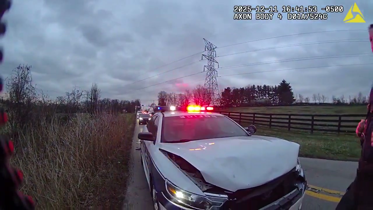Lexington police cruiser with a crumpled hood and emergency lights on, stopped on a two-lane rural road under an overcast sky