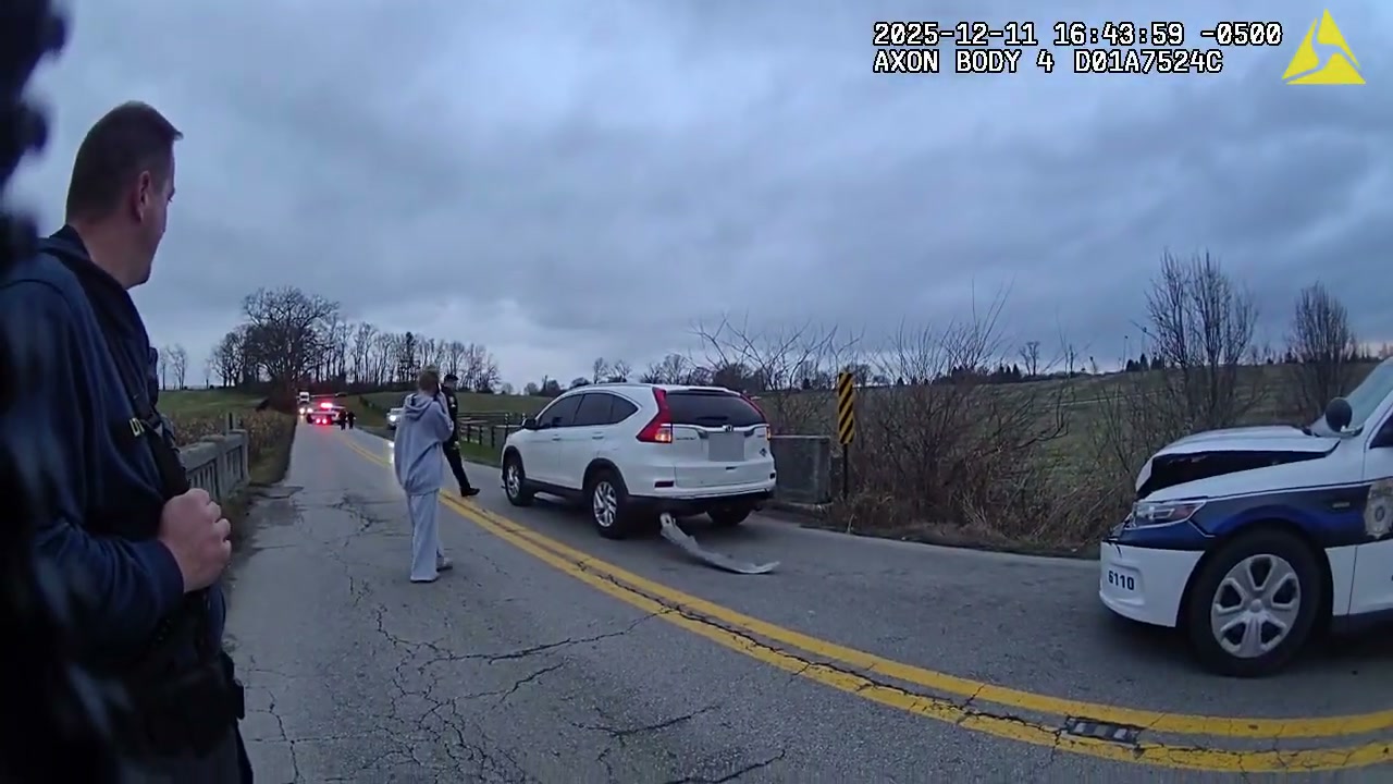 A police supervisor stands on a two-lane road looking toward a white SUV with its rear bumper torn off; a damaged police cruiser sits at right