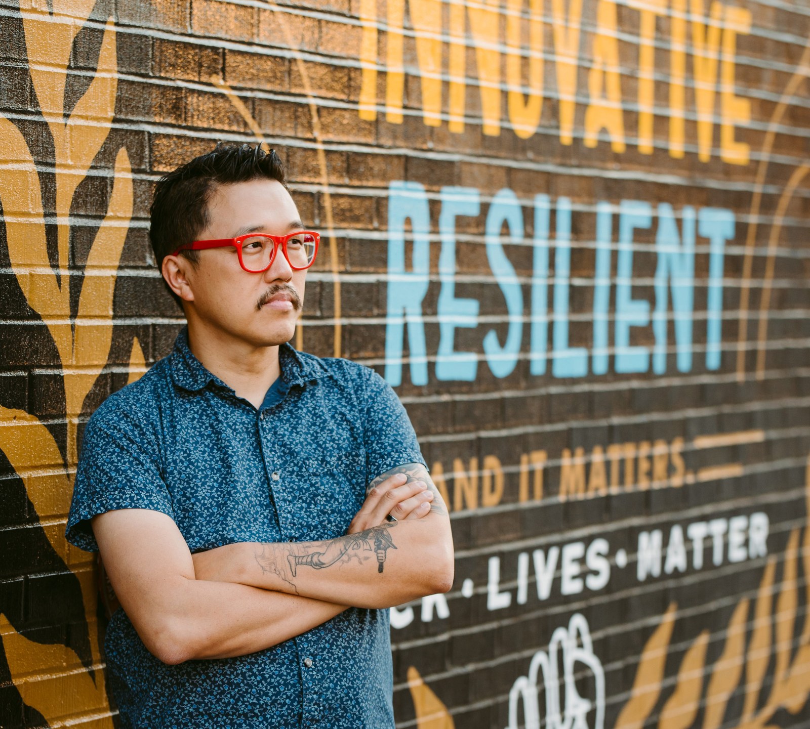 Dan Wu leaning against a brick mural painted with the words Innovative, Resilient, and It Matters, and Lives Matter, arms crossed