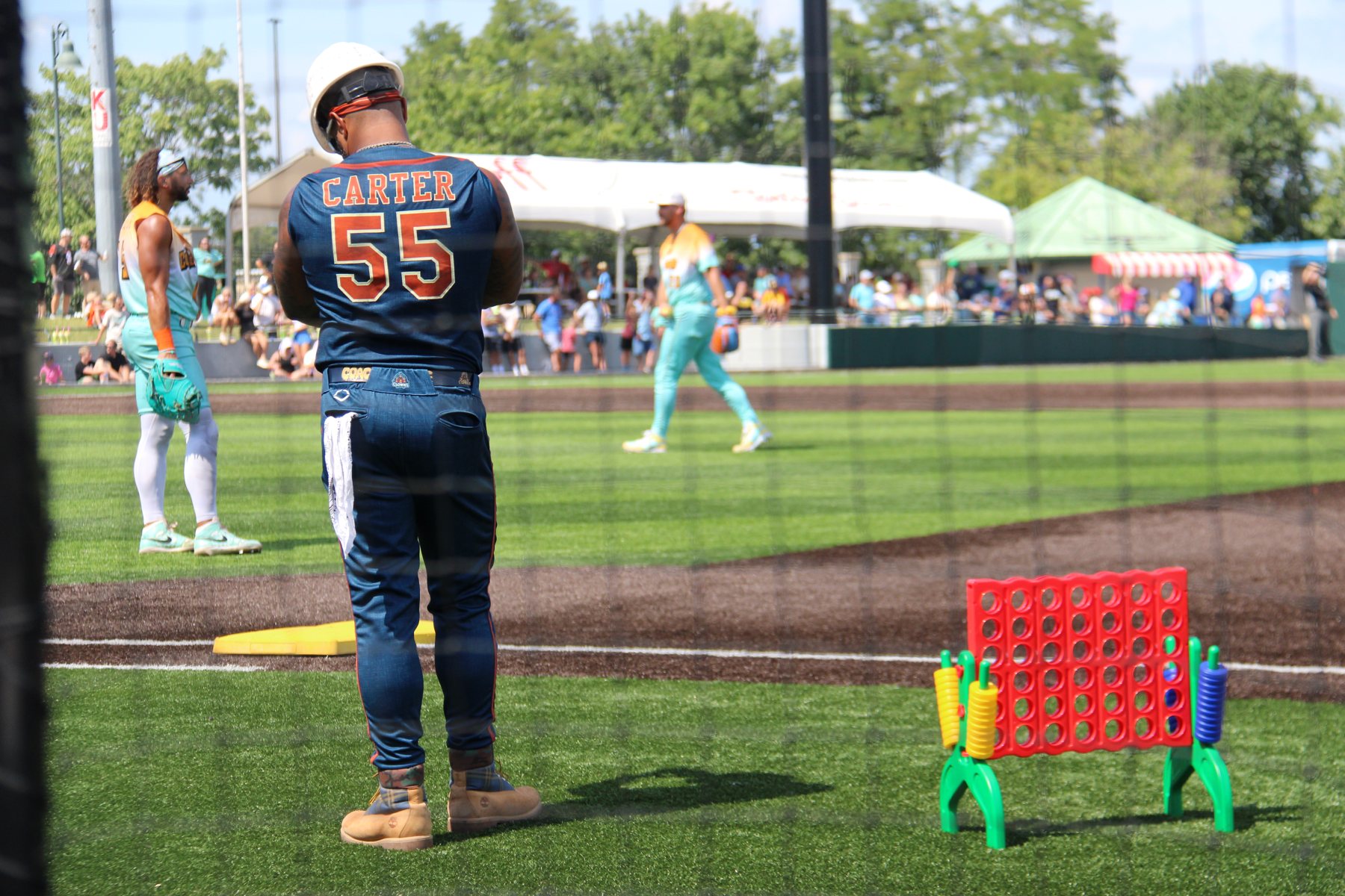 A Clowns coach in a hard hat watches from the first-base coach's box beside a giant Connect Four set.
